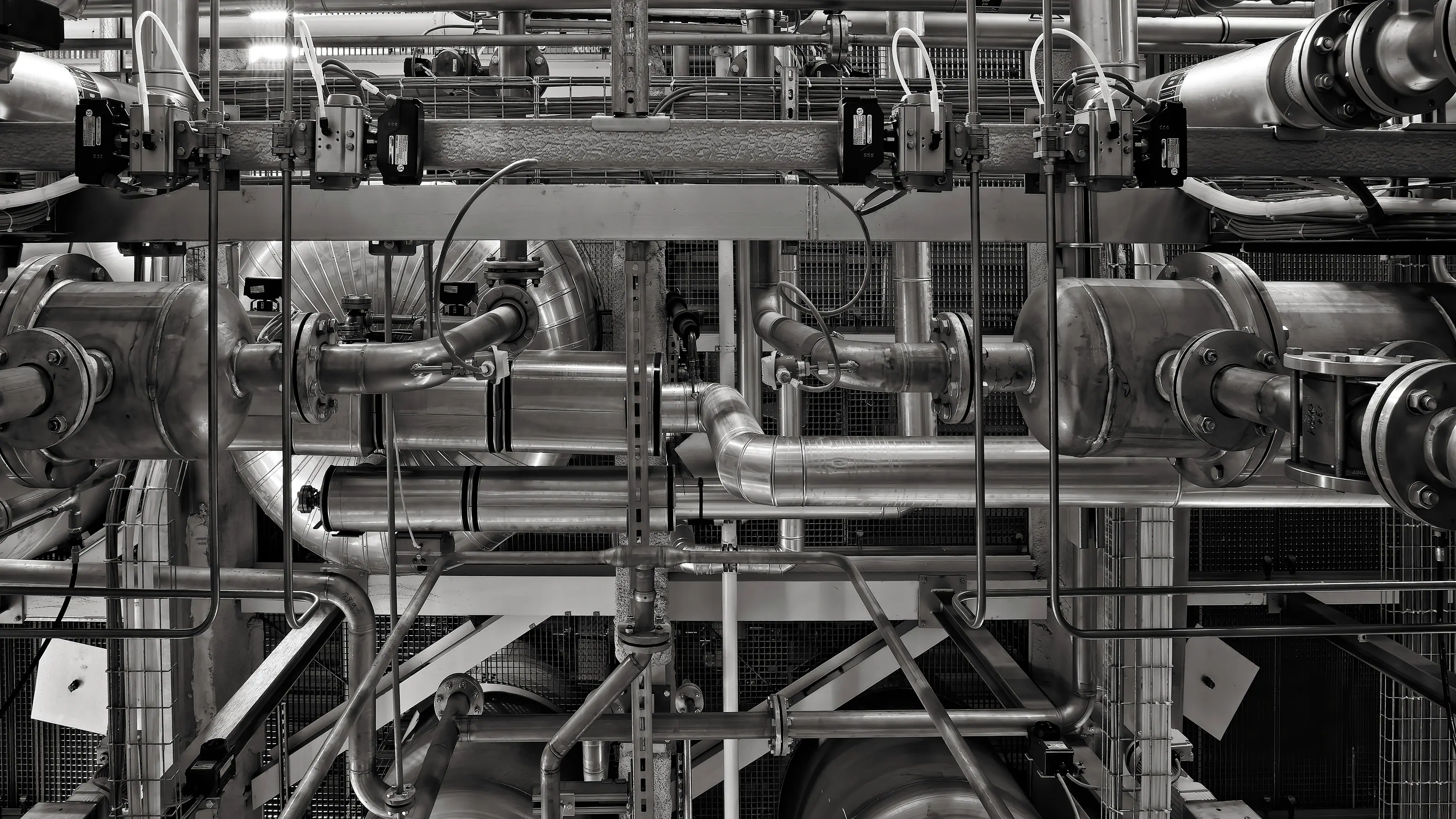 Black-and-white view of stainless steel process pipework, flanged vessels and instrument valves on a steel support frame in a plant room